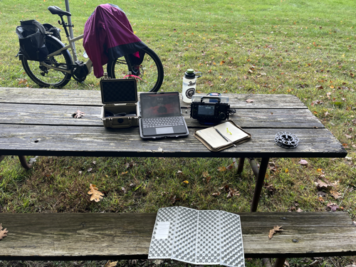 Bike covered with wind shell supporting a mast with computer, radio, notebook, and other gear on a picnic table.