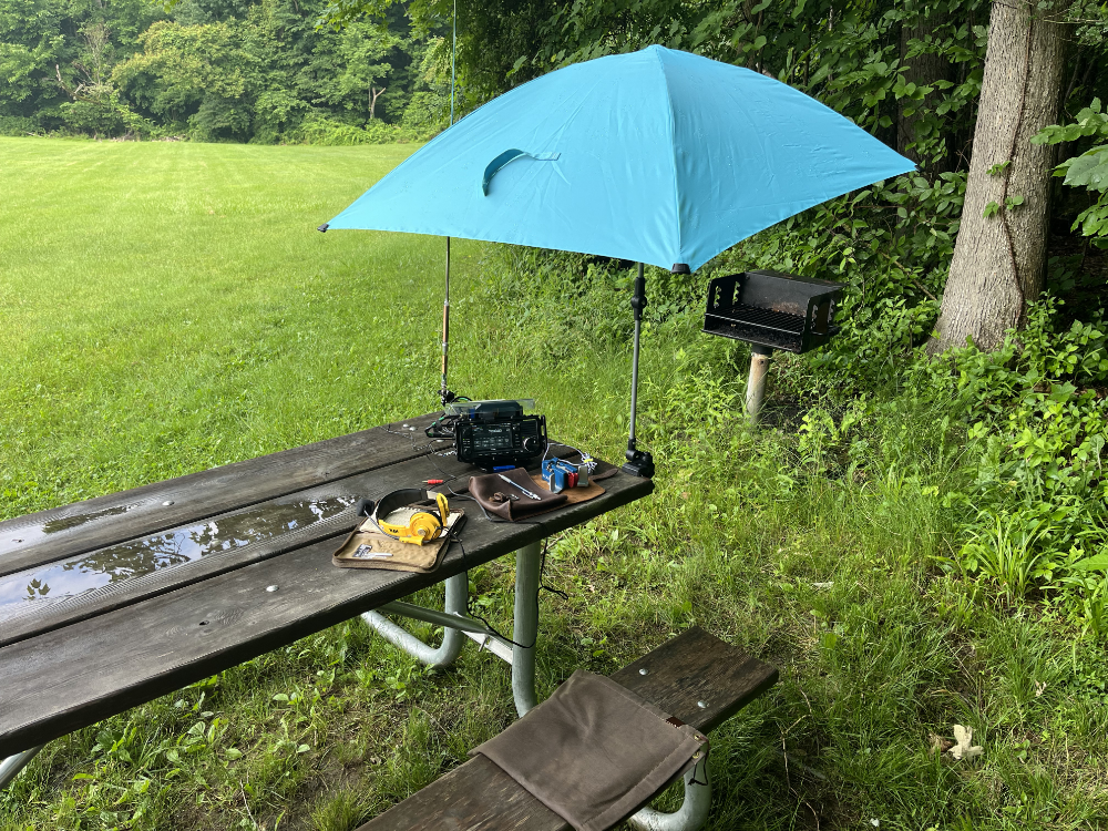 IC-705 and AX1 on a wet table with headphones, log book, and paddles under an umbrella.