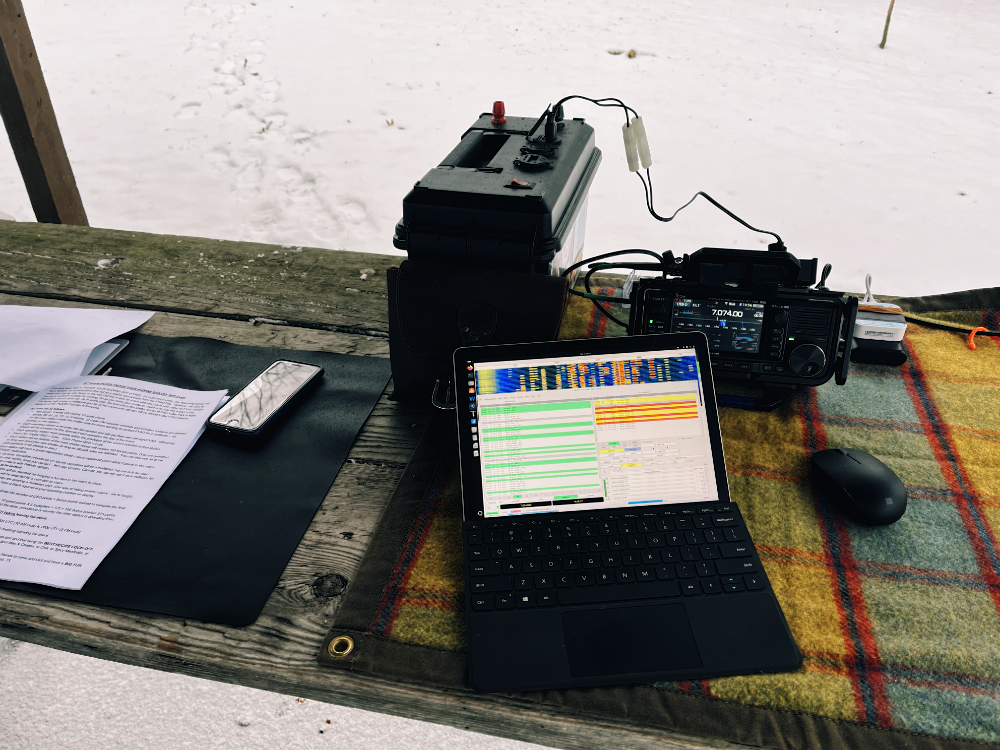 Rules and logging sheets on a picnic table next to a battery box, Surface GO 2, IC-705 and mouse. The setup is clearly too close to the edge of the shelter to keep rain off of it.