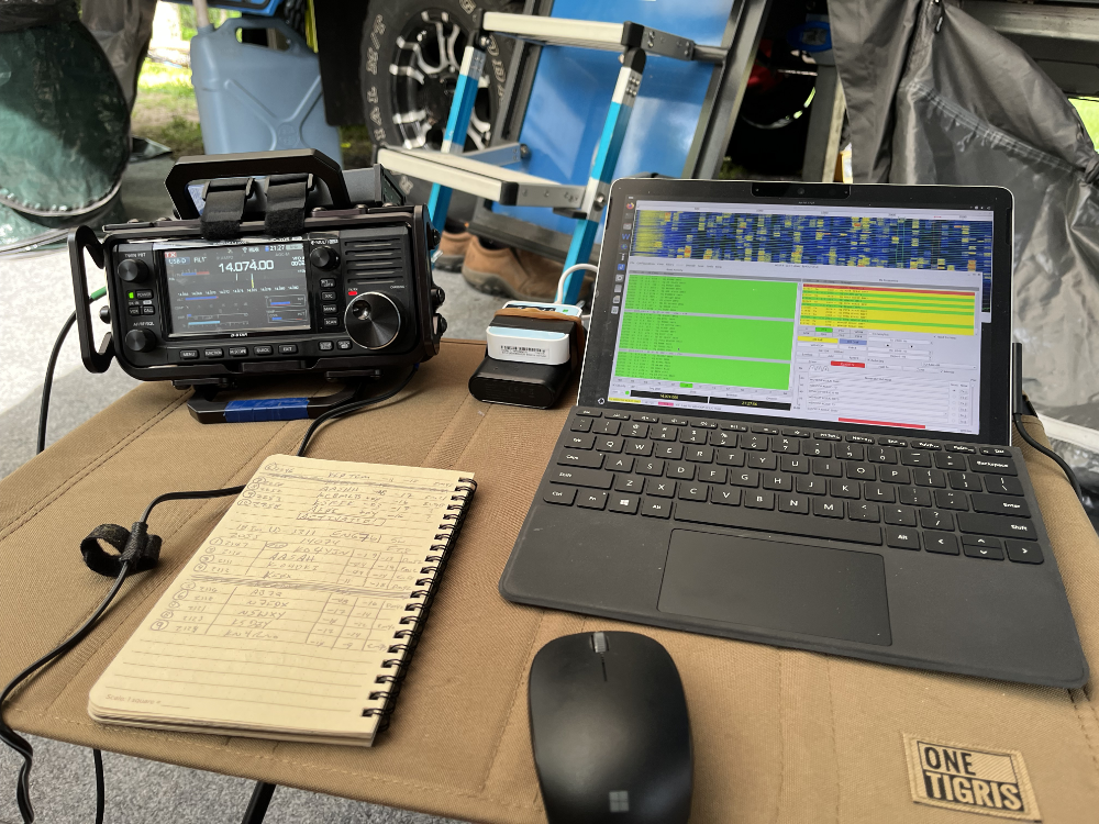IC-705, log book, wireless router with battery pack, Surface GO 2 tablet, and mouse on a folding camp table.