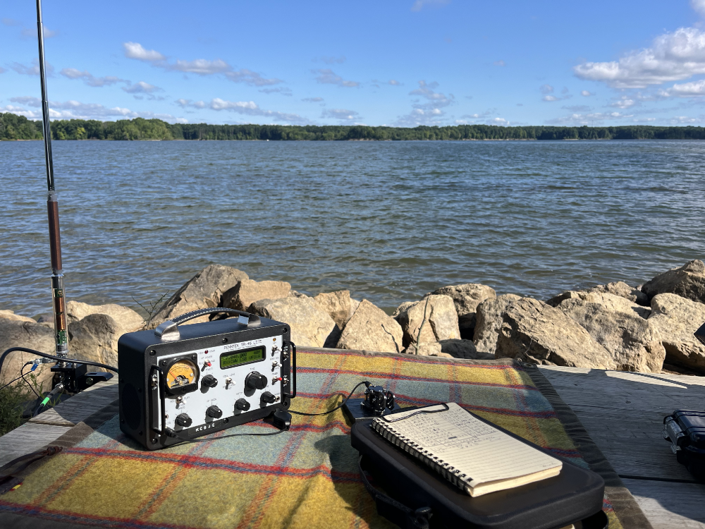 A TR-45L next to a deployed AX1 antenna. There is a set of paddles and a clipboard with log book in the foreground. Everything resting neatly on a gorgeous canvas tarp.
