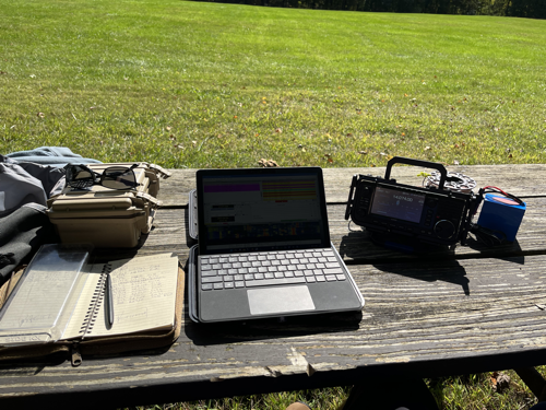 Notebook, Surface Go 2, IC-705 and battery on a picnic table.