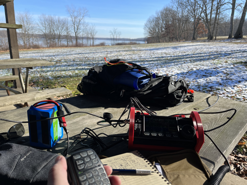 A picnic table with a blue LiFePO battery, log book, and X6100 in the foreground. In the background is a small backpack. My hand is holding the mic.