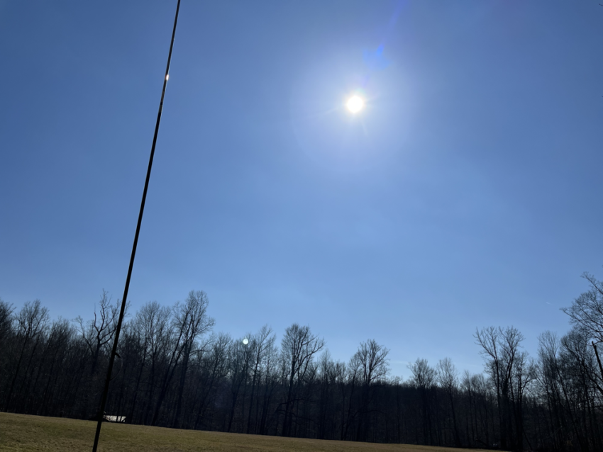 Vertical antenna next to the sun against a blue sky.