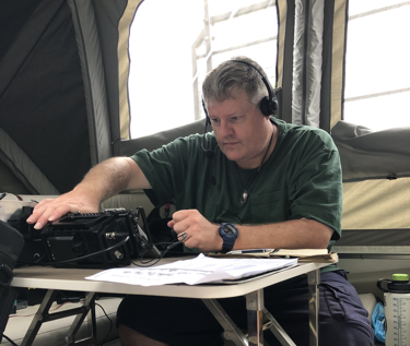A big white guy with grey hair in a green t-shirt operating a radio in a camper.
