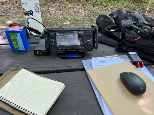 Notebook, IC-705, tuner, battery, water bottle, mouse, paddles and sneaky backpack on a picnic table.