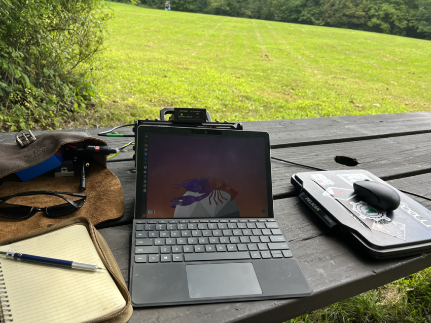 Tablet computer with notebook, pencil, and gear bags on a picnic table.