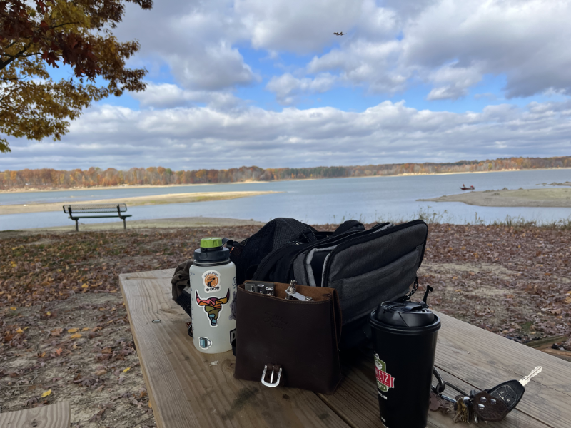 A striking sky with clouds over the reservoir in the background. In the foreground, there is a backpack, water bottle, coffee cup, and a pouch with radio gear inside of it.