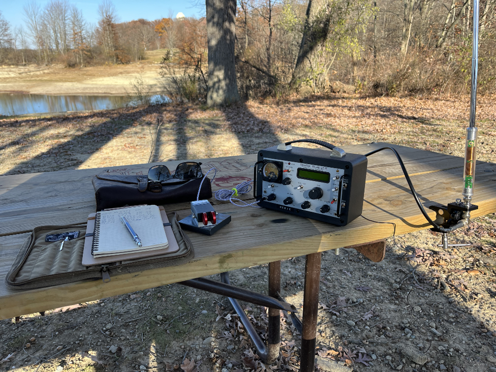 Logbook, CW Morse Paddles, TR-45L, and AX1 on a Standard Issue Park Picnic Table with a leather bag and sunglasses behind them.