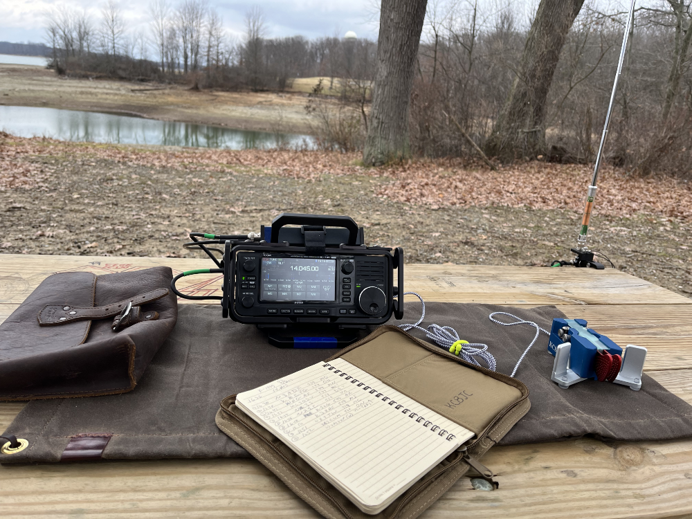Gear on a picnic table: a leather bag, the IC-705, log book, AX1 in the background, and a key resting on a canvas tarp.