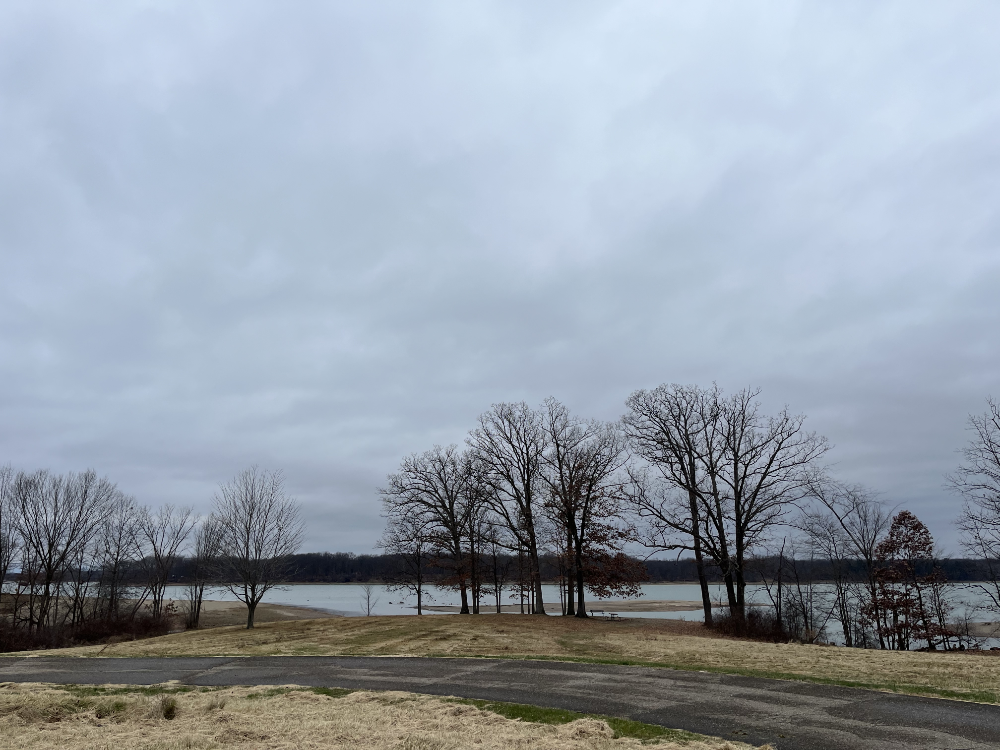 Grey skies over the reservoir. The trees are bare, the grass is brown, and the road around the parking lot is empty. The sun is not present.