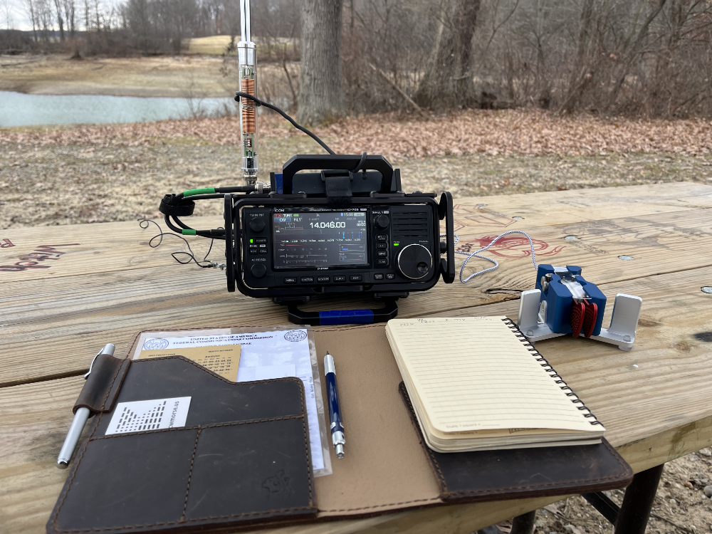 Logbook, IC-705 with tuner and antenna attached next to paddles on a picnic table with water and trees in the background.
