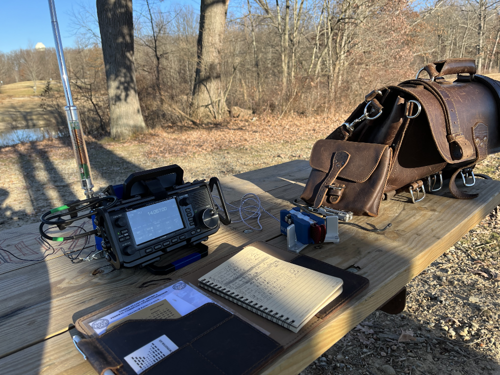 IC-705 with AX1 antenna on a picnic table with log book, Begali Traveller paddles, and leather bags.