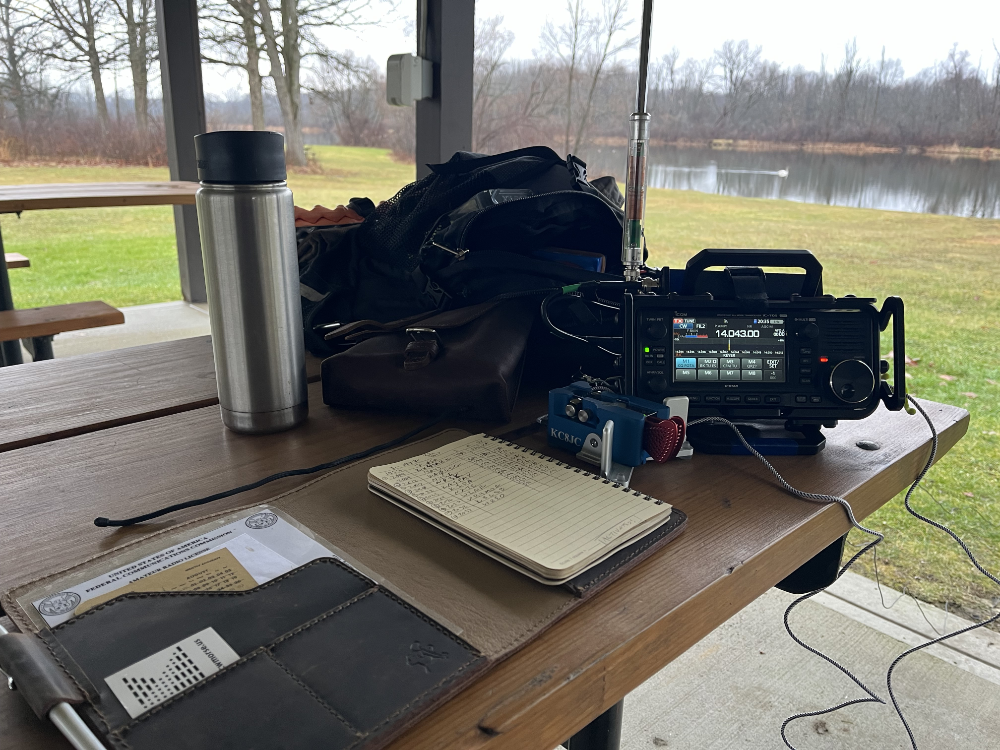Logbook, Begali Traveller Paddles, IC-705 with AX1, coffee cup, and backpack on a picnic table with a pond in the background.