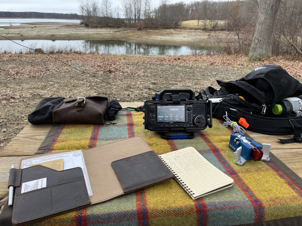 Log book, IC-705, Begali Traveler Key, and assorted bagson a plaid tarp on a picnic table with the reservoir in the background.