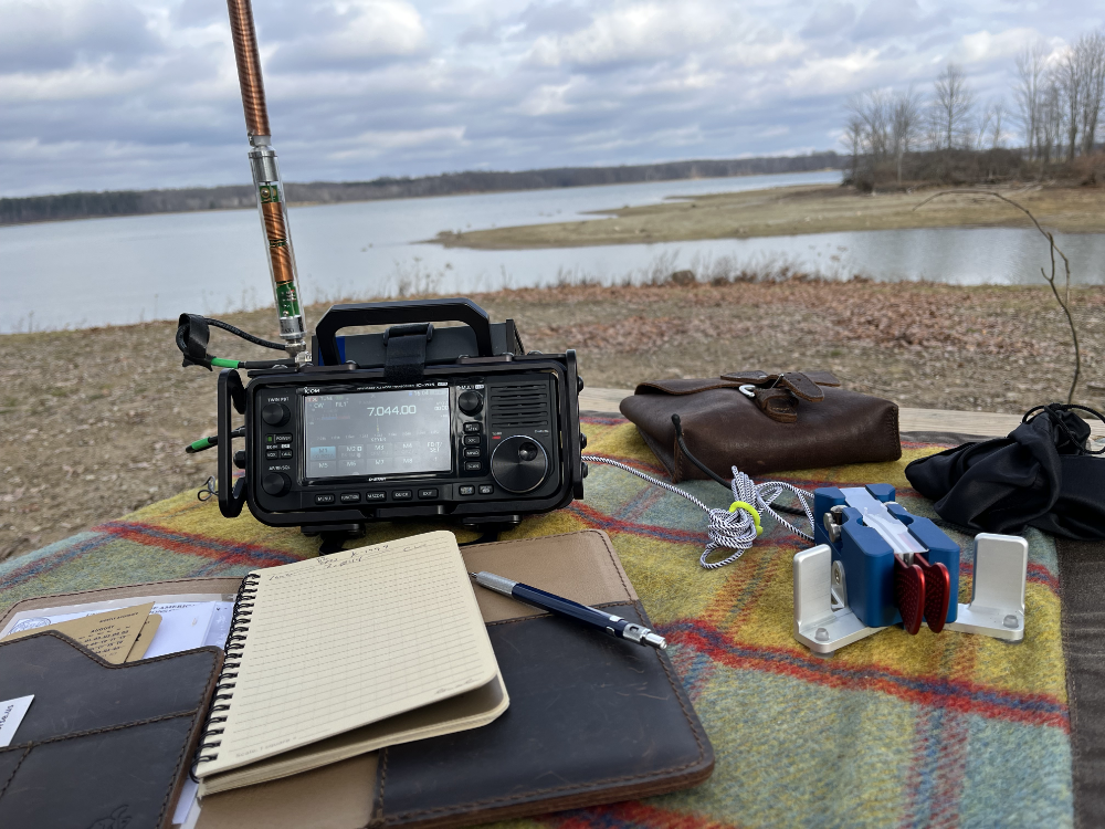 IC-705 with AX1 on a plaid tarp with a log book, gear pouch, and Begali Traveller paddles.