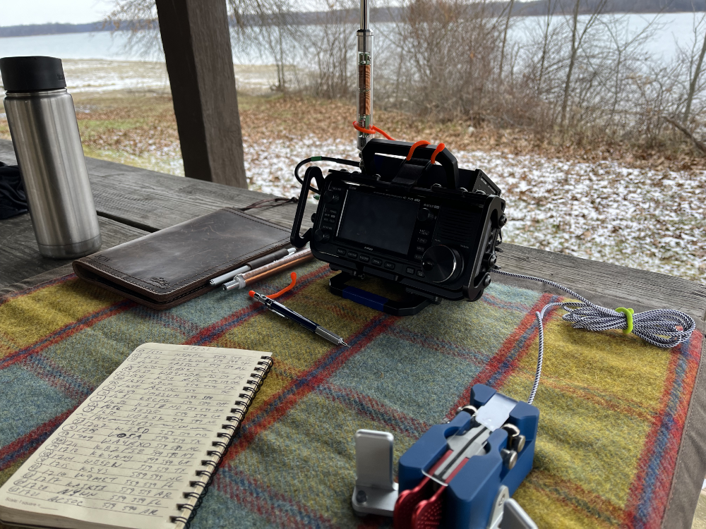 IC-705 on a tarp with logbook, Begali Paddles, and coffee cup. There is a view of the reservoir in the background.