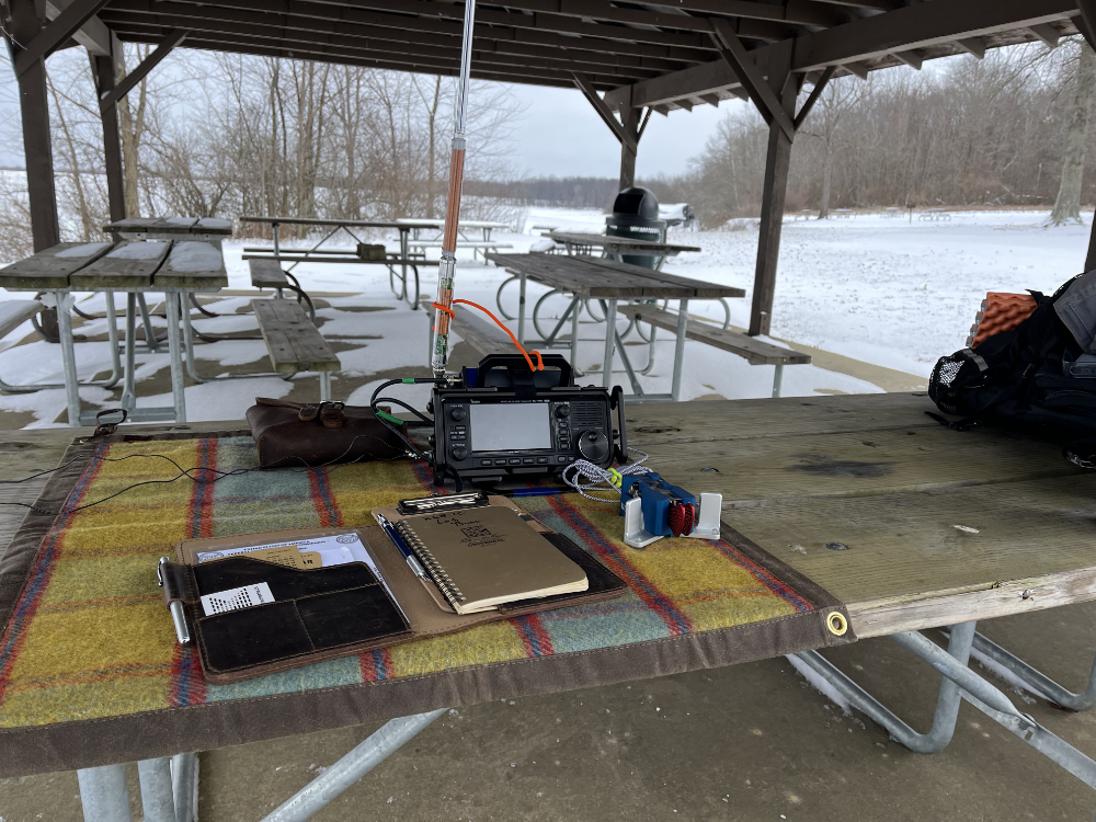 IC-705 on a tarp with a leather pouch, Begali Traveller Light paddles, and log book inside of a picnic shelter looking out on a snowy landscape.