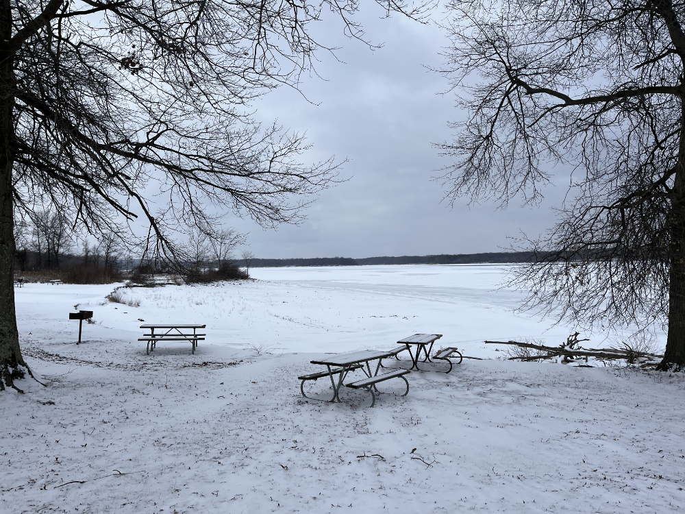 A view of the reservoir with snow covered picnic tables in the foreground. Not a boat to be seen on the frozen water.