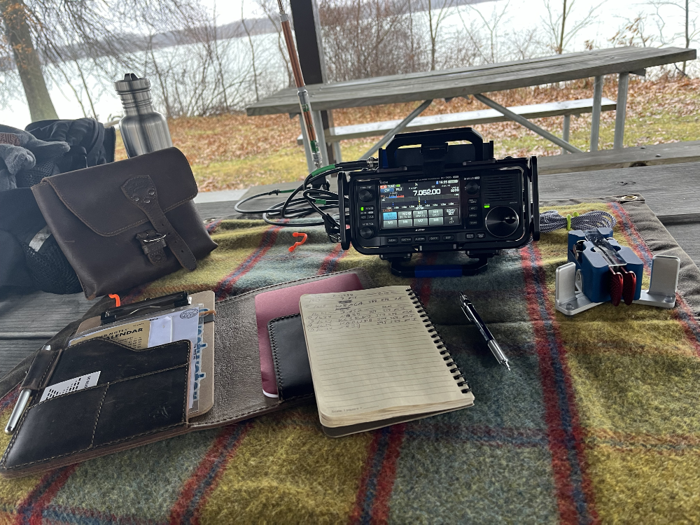 IC-705 on a tarp sitting on a picnic table next to a Begali Traveler key and a log book.