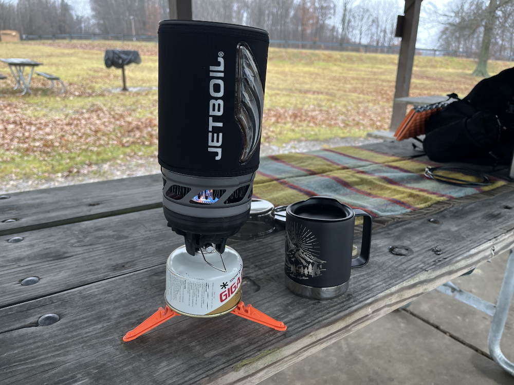 JetBoil container on a burner boiling water next to a coffee cup on a picnic table.