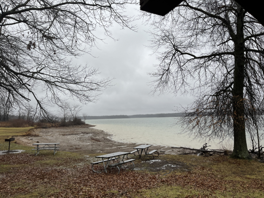 Picnic tables on a piece of land close to the shore of a reservoir in the rain under a grey sky.