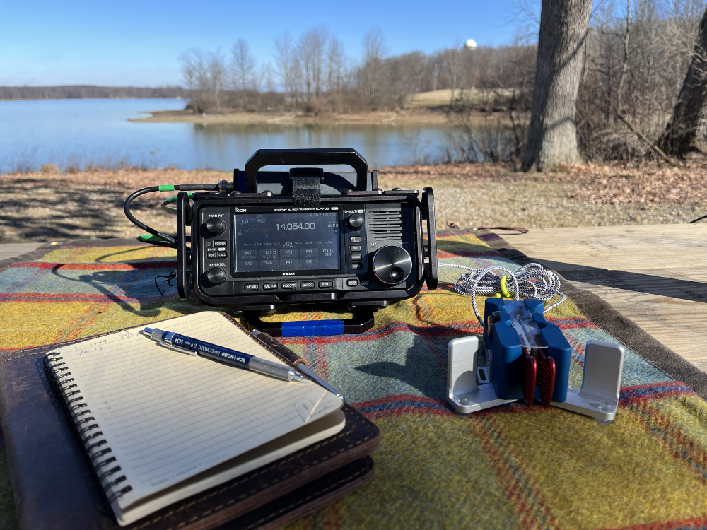 IC-705, Begali Traveler, and log book resting on a tarp on a picnic table with the reservoir in the background.