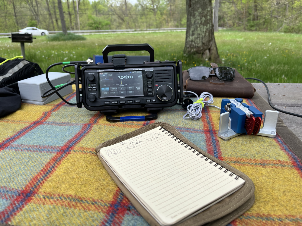 IC-705 and Begali Traveler on a tarp with a notebook in the foreground and assorted containers on the periphery.