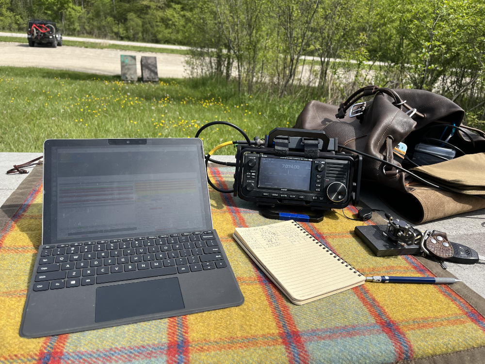 Surface Go 2, IC-705, log book, pencil, Putikeeg paddles, and backpack on a tarp. The Jeep and fat bike are in the parking lot beyond.